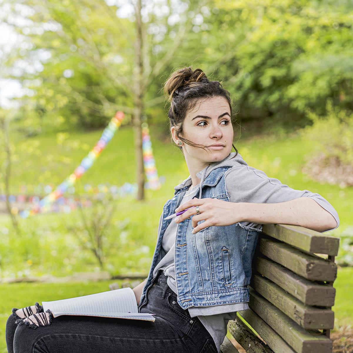 Photo of a female Chatham student with a pencil in hand, seated on a bench outside on Shadyside Campus with an open notebook in her lap